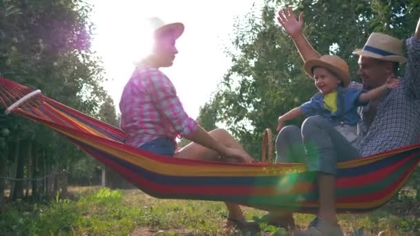 Maison de vacances, les parents heureux avec l'enfant sur l'hamac dans le jardin de pomme s'amusent et apprécient la nature dans des chapeaux de paille à la chaude journée d'été — Video