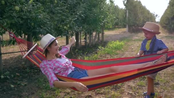 Vacanță în grădină de mere, copil fericit leagănă mama lui într-un hamac între rânduri de copaci — Videoclip de stoc