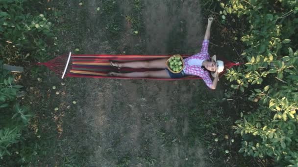 Vue sur le jardin, jeune femme mange une pomme et se détendre dans un hamac avec panier de fruits — Video