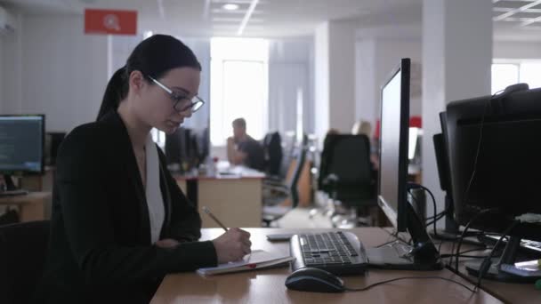 Špatná práce, zamračená dívka s brýlemi pracuje v kanceláři u počítače a dělá si poznámky do zápisníku — Stock video