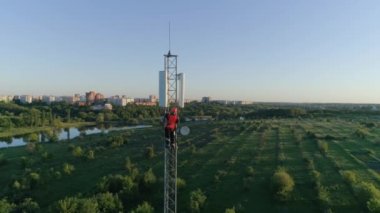 Anten iletişim kulesi, insanın hava görüntüsü günbatımının arka planındaki güvenlik ekipmanlarını kullanarak yükseklerde çalışıyor.