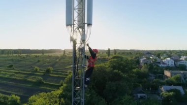 Güvenlik yeleği ve başlık mühendisi, telekomünikasyon kulesinin insansız hava aracı görüntüsü olan hücresel antenin tepesine tırmanıyor.