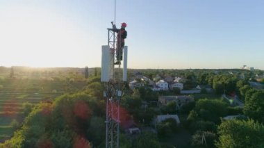 Üst hücre antenindeki teknisyen mavi gökyüzünün ve güneş ışığının arka planında telekomünikasyon kulesinin hava görüntüsünü gösteriyor.