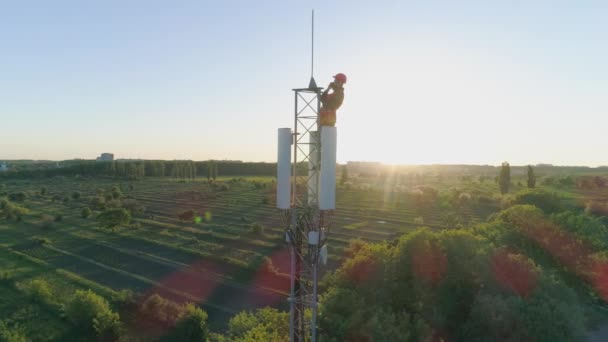 Wieża komunikacyjna z wykonawcą sprawdzająca połączenie mobilne na szczycie anteny komórkowej na tle błękitnego nieba z zachodem słońca — Wideo stockowe