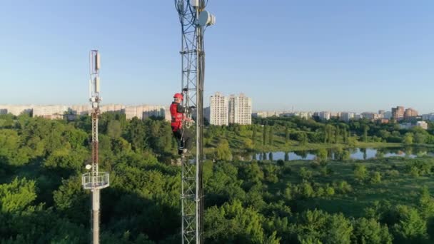 Pracovník v hard hat stoupá vysoko na rádiové telekomunikační věž na pozadí krásné městské krajiny, letecký pohled na mobilní antény — Stock video