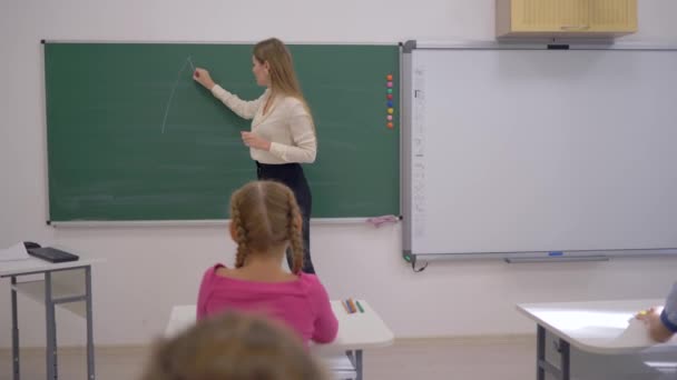 Junior School, nauczycielka rysuje geometryczne kształty na pokładzie podczas lekcji dla uczniów siedzących przy biurkach w klasie — Wideo stockowe