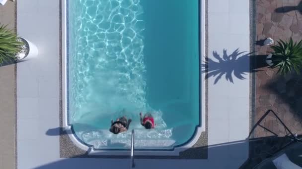 Беспилотник смотрит на длинноволосых девушек в купальниках плавающих в голубом бассейне на летнем курорте — стоковое видео