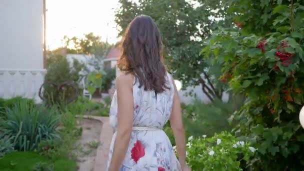 Fascino femminile, dolce ragazza in abito volante passeggiate e si guarda intorno nel cortile tra gli alberi — Video Stock