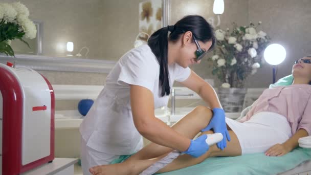 Péče o pleť, profesionální kosmetolog do lékařských brýlí dělá fotoepilaci na nohách paní klientky pomocí laserového stroje v beauty studiu — Stock video