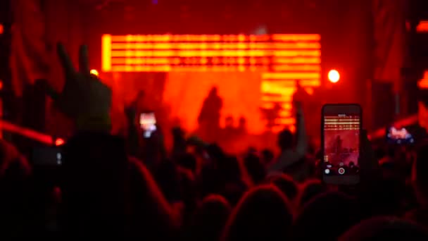 Многие размахивают руками и записывают видео на смартфон рок-группы на сцене в подсветке прожекторов — стоковое видео