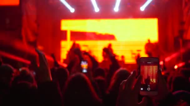 Рок-вечеринка, много людей размахивают руками и записывают на видео выступления музыкальных исполнителей на сцене при прожекторе — стоковое видео