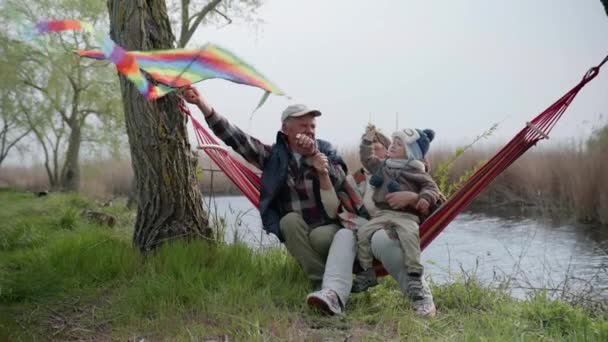Family, joyful little grandson is sitting on hands of caring and joyful grandad loving grandmother in hammock and playing with kite — Stock Video