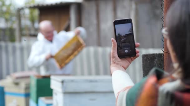 Пожилой пчеловод принимает видео контент на мобильный телефон для блога с помощью жены и показывает подписчикам соты — стоковое видео