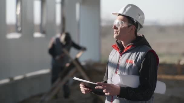 Sorridente ingegnere maschio in casco protettivo con disegni in mano, sorride e ispeziona la costruzione di un nuovo hangar — Video Stock
