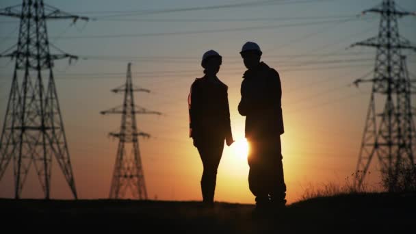 Silhouet, mannelijke en vrouwelijke ingenieurs in beschermende helmen gebaren met hun handen, bespreken de bouw van een nieuwe elektriciteitsleiding — Stockvideo