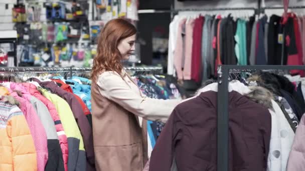 Shopaholic, mujer joven atractiva que intenta en ropa de abrigo hermosa en tienda de moda durante la venta en el centro comercial — Vídeos de Stock