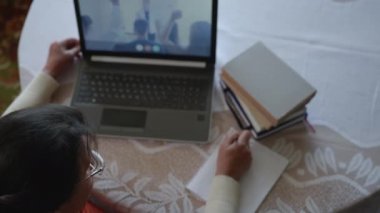 Üst görüşe göre, yaşlanan kadın, online eğitim sırasında daha fazla öğrenim görüyor, elini kaldırıyor ve cevapları kazı kazan ile yazıyor.