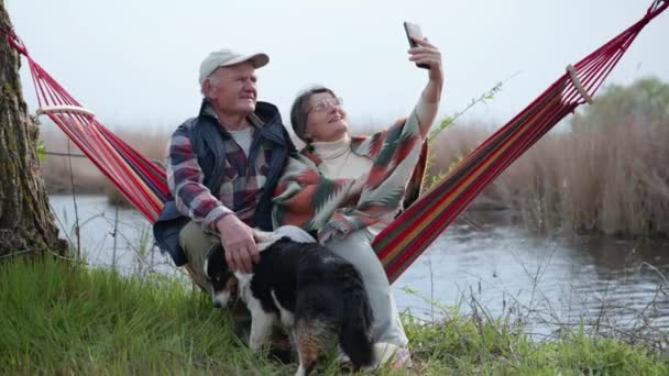 Loving elderly couple cheerful man and woman sitting together in hammock by river and talking with their dear children using modern technology using smartphone — Stock Video