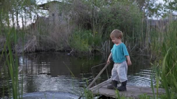 Детство, маленький счастливый мальчик в резиновых сапогах играет на пирсе возле реки с палкой — стоковое видео