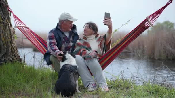 Glad äldre man och hustru ha kul sitter i en hängmatta vid floden med en hund helvete som en prod och tillbringa tid tillsammans prata på en mobiltelefon — Stockvideo