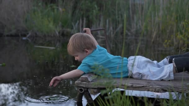 adorable garçon se trouve sur la jetée près de l'étang et joue dans l'eau avec des feuilles, petit enfant joue près de l'eau fond de roseaux 