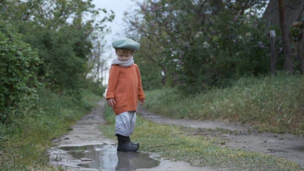 Счастливое детство, привлекательный мальчик в шляпе и резиновых сапогах, развлекаясь, играя на свежем воздухе, прыгая в лужу на дороге после дождя — стоковое видео