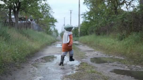 Feliz infancia, chico divertido en sombrero y botas de goma disfruta de caminar por la calle, corre divertido a través de charcos después de la lluvia — Vídeos de Stock