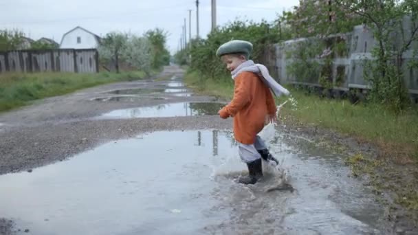 面白いゲーム、帽子とゴムブーツの魅力的な陽気な少年は暖かい春の日に雨の後、通りの水たまりで遊ぶ — ストック動画