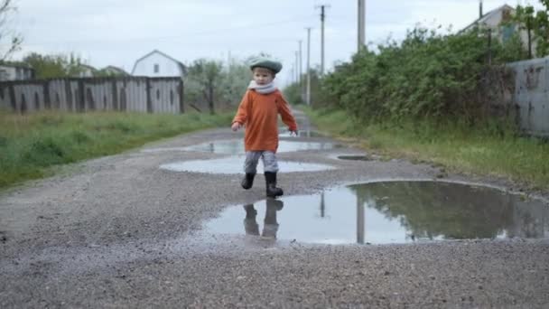 Szabadtéri játékok, szerethető vidám fiú kalap és gumicsizma játszik a pocsolyák az utcán eső után — Stock videók