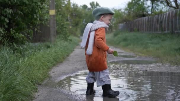 Juegos al aire libre, niño feliz en sombrero y botas disfruta jugando al aire libre y lanza hojas en el charco — Vídeos de Stock