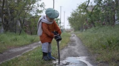 Yağmur üstüne eğlence, neşeli çocuk sokakta oynamaktan hoşlanıyor, lastik botlarıyla oynamaktan zevk alıyor, çocuk bir su birikintisinden botuna su çekip geri dolduruyor.