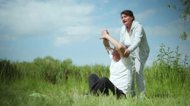 Yoga och meditation, vårdande gammal fru hjälper sin älskade äldre man utföra kroppsövningar medan hon sitter på yogamattan under morgonövningar utomhus — Stockvideo