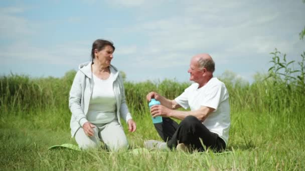 Cura uomo anziano dà acqua fresca e pulita in bottiglie di nutrizione sportiva alla sua cara moglie amata dopo aver praticato sport o meditare all'aperto — Video Stock