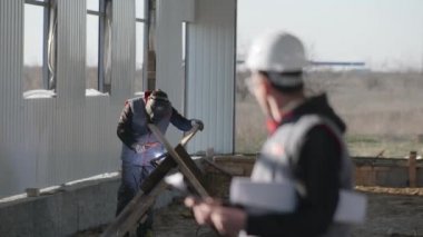 Bina, koruyucu kasklı ve ellerinde çizimler olan yakışıklı erkek mühendis, gülümsüyor ve yeni hangarın inşaatını denetliyor.