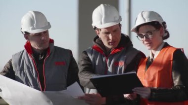 Koruyucu miğferli mühendis ve mimarlardan oluşan dost bir ekip, inşaat alanında çizimleri tartışıyor.
