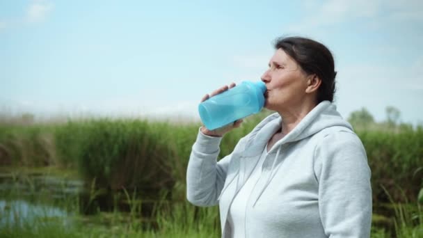 Mulher muito velha sacia sede bebe água depois de fazer esportes ao ar livre no prado por rio — Vídeo de Stock