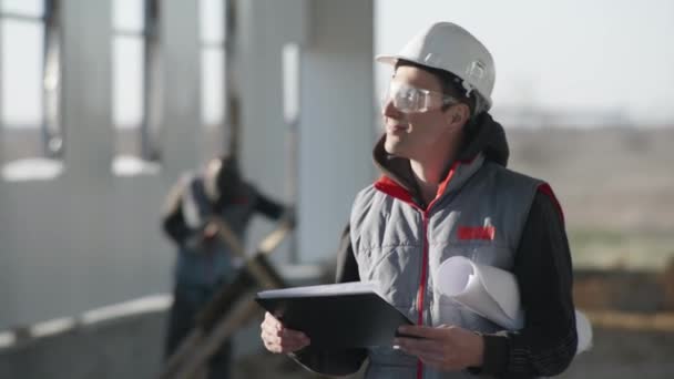 Ingegnere professionista sorridente in uniforme protettiva con occhiali di sicurezza e casco sullo sfondo di un saldatore e scintille di saldatura durante la costruzione di un grande impianto industriale — Video Stock