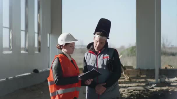 Engenheira industrial feminina em capacete protetor discute novo projeto com desenhos em mãos com uma funcionária em equipamentos de trabalho e capacete durante construção de hangar — Vídeo de Stock