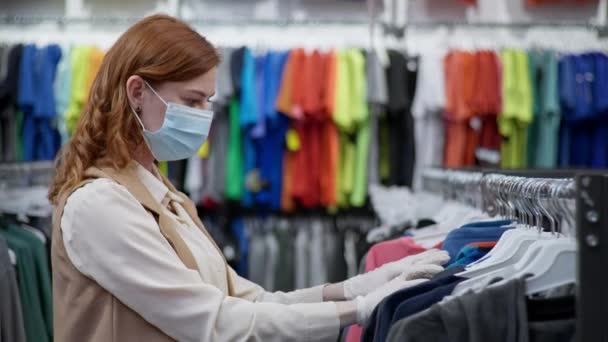 Pandemie, hübsches Käufermädchen in medizinischer Maske und Handschuhen wählt Kleidung in den Regalen — Stockvideo