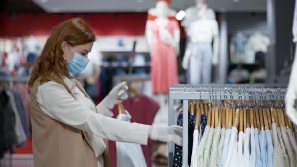 Minorista, comprador bastante femenino en una máscara médica y guantes compra ropa de moda en los estantes de la tienda — Vídeos de Stock