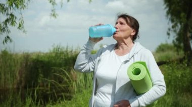 Yoga ya da meditasyon için halısı olan neşeli, yaşlı bir kadın. Sağlığını korumak ve temiz su içmek için ellerinde.