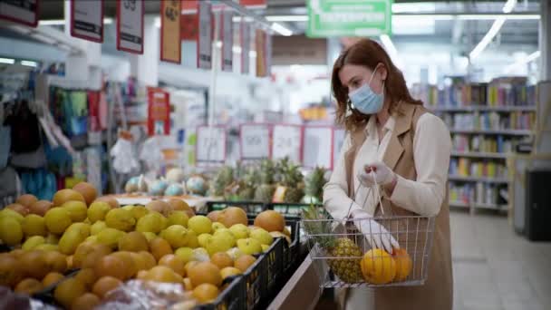 Toko gadis bertopeng dan sarung tangan untuk melindungi dari coronavirus dengan keranjang makanan di tangannya membeli buah-buahan yang sehat untuk mendukung kekebalan selama pandemi karantina di supermarket — Stok Video