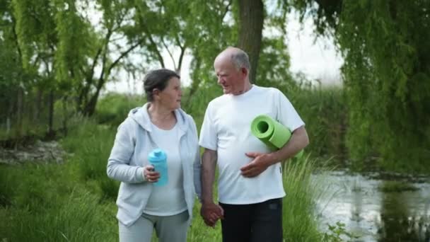 Pareja de ancianos casados con botella de agua para la nutrición deportiva y esterilla de yoga van de la mano después de practicar deportes y ejercicios matutinos — Vídeo de stock