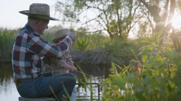 Rybáři, starostlivý milující dědeček spolu s krásným šťastným vnukem, rybaření pomocí provizorního rybářského prutu sedí na molu u řeky v teplém odpoledni — Stock video