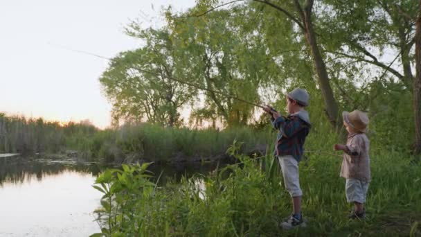 Ländliches Wochenende, glückliche unbeschwerte Jungen genießen die Natur und angeln mit der Angelrute am Flussufer zwischen Bäumen und untergehender Sonne — Stockvideo