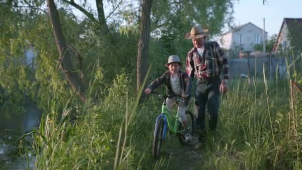 Little boy in a hat learns to ride a bicycle with the help of his grandfather — Stock Video