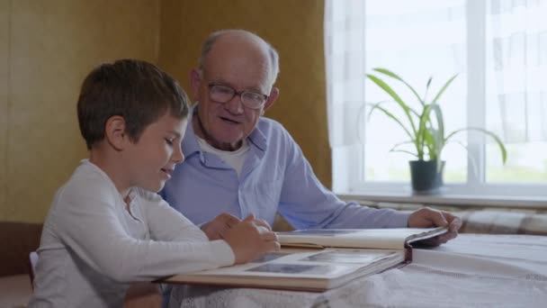 Recuerdos del hogar, pequeño niño atractivo con su abuelo de edad avanzada divertirse mirando un álbum de fotos — Vídeo de stock