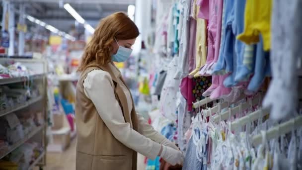 Atractiva mujer feliz en una máscara y guantes para proteger contra la infección por virus o coronavirus compra ropa para niños en los estantes de la tienda al por menor — Vídeos de Stock