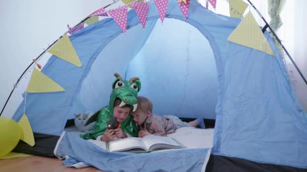 Lazer das crianças, belos meninos amigáveis em um traje de dinossauro se divertir lendo um livro e jogando um smartphone com seu irmãozinho bonito em um wigwam — Vídeo de Stock