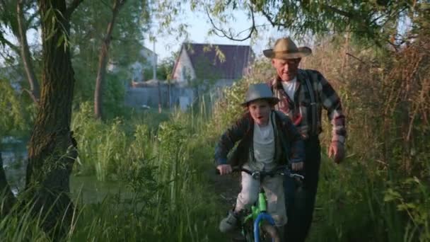 Schattig mannelijk kind in een hoed leert fietsen met behulp van zijn opa achtergrond van het meer op het platteland — Stockvideo